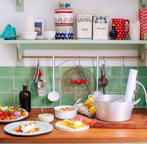 A styled kitchen scene with a wooden countertop holding plated food, a silver pot, a white electric masher, hanging utensils, and decorative canisters on a shelf above a green tiled backsplash.